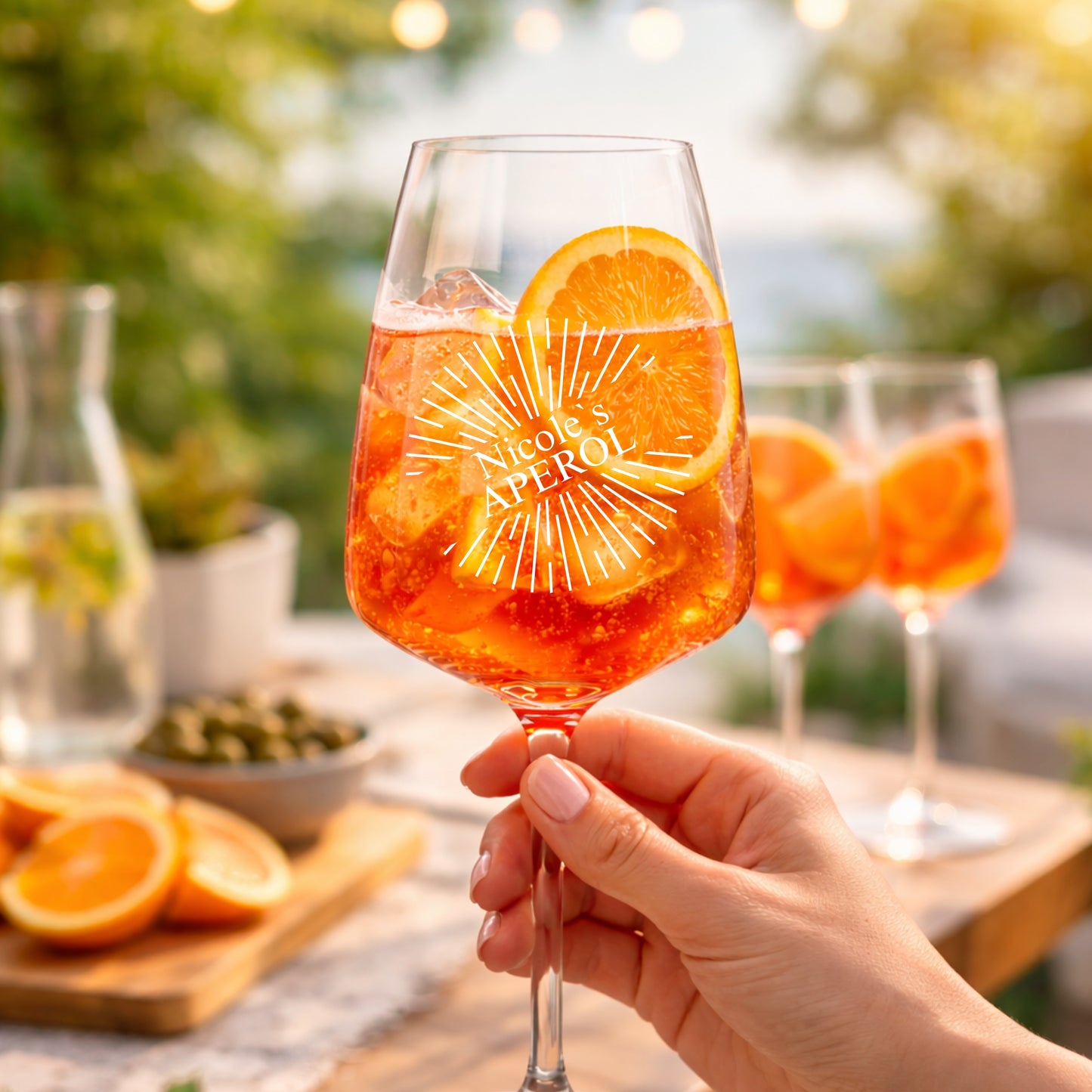 Hand hält personalisiertes Aperol Spritz Glas mit Sunburst-Gravur und Namen Nicole – sommerliche Gartenparty